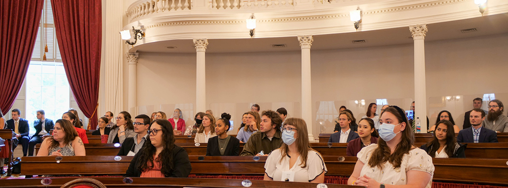 A diverse group of Law Students attends an event at the Vermont State Capitol Building, sitting at the desks of legilstators